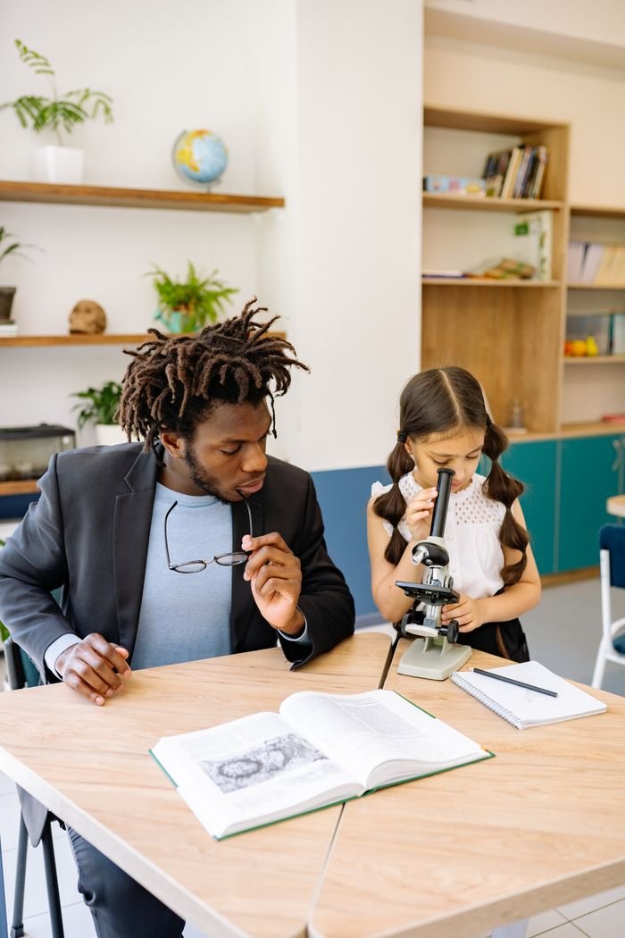 About A teacher helps a young student use a microscope in a classroom setting.